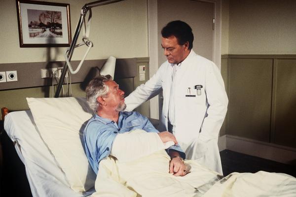 Bild 1 von 1: Der Patient Willi Anschütz (Klaus Havenstein, l.) soll am Blinddarm operiert werden. Aber je näher der Termin rückt, desto mulmiger wird ihm ums Herz. Sein Horoskop empfiehlt ihm, kein Risiko einzugehen. Prof. Brinkmann (Klausjürgen Wussow) versucht ihn zu überzeugen, dass er die Operation dringend benötigt.