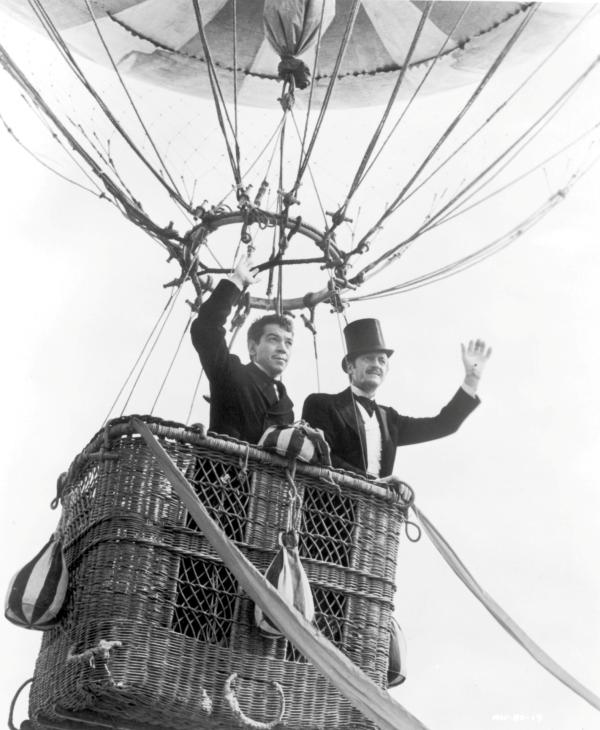Bild 1 von 11: Full shot of Cantinflas as Passepartout and David Niven as Phileas Fogg, wearing top hat; both standing and waving in basket of hot air balloon.