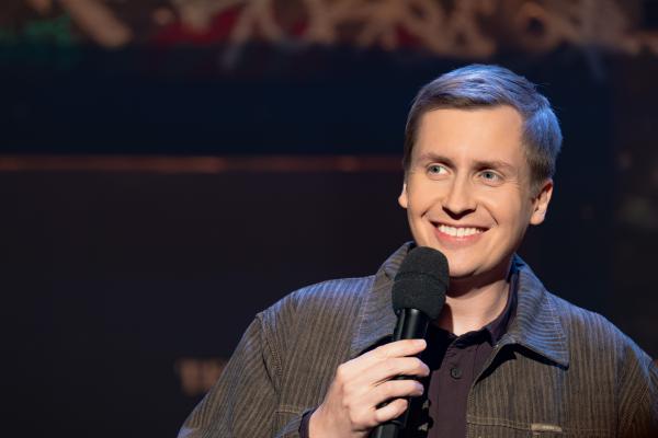 Bild 1 von 9: Stand-Up-Comedian und Gastgeber Till Reiners steht mit einem Mikrofon in der Hand auf der Bühne und spricht zum Publikum.