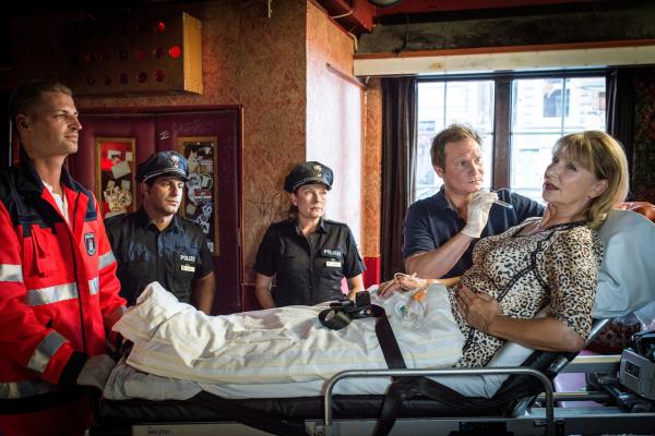 Bild 1 von 11: Dr. Haase (Fabian Harloff, 2.v.r.) untersucht Ingrid Ruhland (Sabine Kaack, r.), die bei dem Überfall auf ihre Bar mit einem Baseballschläger verletzt wurde. Claudia (Janette Rauch, M.) und Tarik (Serhat Cokgezen 2.v.l.) warten mit ihrer Befragung, bis Haase fertig ist.