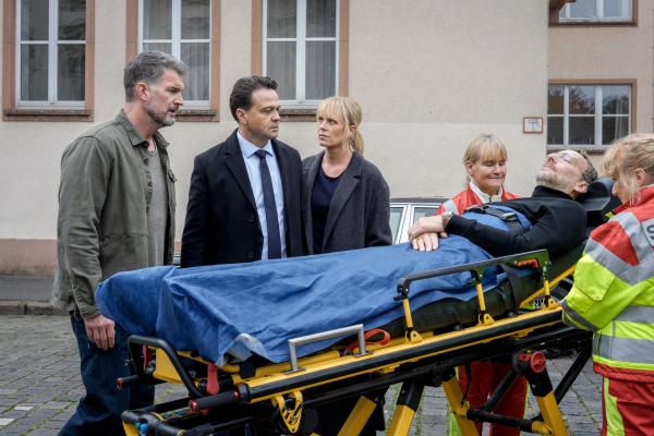 Bild 1 von 4: Jemand scheint es auf den Bekanntenkreis von Staatsanwalt Binz (Michael Rotschopf, 2.v.l.) abzusehen. Gemeinsam mit Dirk Trebbe (Simon Böer, l.) und Ina Zimmermann (Melanie Marschke, M.) beobachtet er, wie Kilian Cornelsen (Knut Berger, 2.v.r.) von Sanitäterinnen (Komparsinnen) weggefahren wird.
