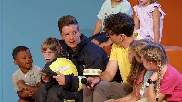 Bild 1 von 5: Im Bild: Moderator Christoph Hirschler und sein Gast, Feuerwehrfrau Katja, sitzen umgeben von neugierigen Kindern und beantworten deren Fragen rund um das Thema Feuerwehr. Die Kinder lauschen gespannt und stellen interessiert ihre Fragen, während Katja Einblicke in ihren spannenden Alltag als Feuerwehrfrau gibt.