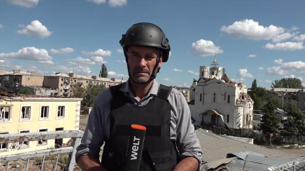 Bild 1 von 3: WELT-Reporter Steffen Schwarzkopf berichtet in Schutzweste und Helm aus einer stark zerstörten Stadt, hinter ihm die Überreste einer Kirche und zerbombter Wohnhäuser.