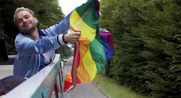 Ein Mann hält eine Regenbogenflagge in der Fahrtwind. Er steht auf einem Fahrzeug.