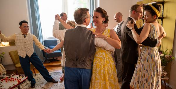 Bild 1 von 3: Während das Ehepaar Ina (Josephine Ehlert, 4.v.r.) und Georg Trapp (Dominik Klingberg, 4.v.l.) eine ausgelassene 50er-Jahre Party feiert, kommt es in dem Mehrfamilienhaus zu einer grausamen Tat: Am Fuß der Kellertreppe wird eine Frau tot aufgefunden - sie wurde gestoßen. Doch weder die Trapps, noch die Gäste (Komparsen) wollen etwas mitbekommen haben.
