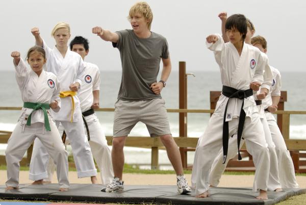 Bild 1 von 7: Lewis (Mitte, Angus McLaren) ist fest davon überzeugt, dass seine Beziehung zu Cleo deshalb gescheitert ist, weil er nicht männlich genug sei und versucht sich daher in Karate.
