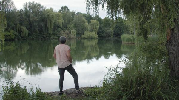 Bild 1 von 7: Protagonistin Angelika steht gedankenversunken an einem See.