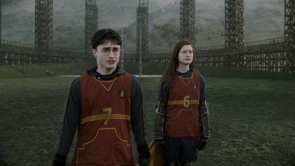 Bild 1 von 15: Daniel Radcliffe as Harry Potter standing in stadium field with Bonnie Wright as Ginny Weasley, both wearing Hogwarts quidditch team uniforms.