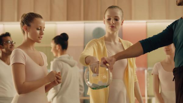 Bild 1 von 6: Für die heutige Aufgabe im Ballettunterricht sollen Paare gebildet werden. Jeder darf eine Zettel mit dem Namen des Tanzpartners ziehen. Thea (Hanna Dodd, M.) fischt gerade einen Zettel aus dem Glas, das Lehrer Armando Castillo (Rik Young, r.) hält, während Lena (Jessica Lord, l.) sehr gespannt darauf wartet, an der Reihe zu sein.