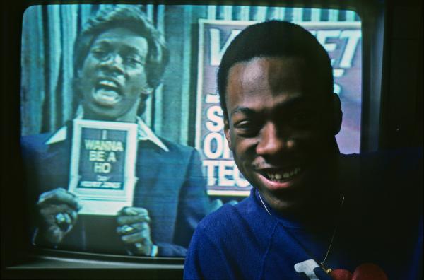 Bild 1 von 1: Eddie Murphy posiert 1982 vor einem Fernseher, der ein Bild seiner Figur Velvet Jones zeigt, die er eigens für ?Saturday Night Live? schuf.