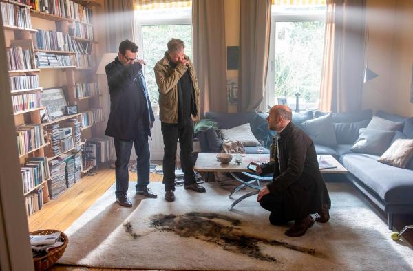 Bild 1 von 7: Im Bild: Die Kommissare Finn (Sven Martinek, l.) und Lars (Ingo Naujoks, 2.v.l.) stehen am Fundort der Leiche von Hans Vonnsen. Auf dem Teppich vor ihnen ist ein dunkler Abdruck des bereits stark verwesten Körpers zu sehen. Dr. Henning Strahl (Christoph Tomanek, r.) kniet auf dem Boden und dokumentiert den Tatort mit seiner Kamera.