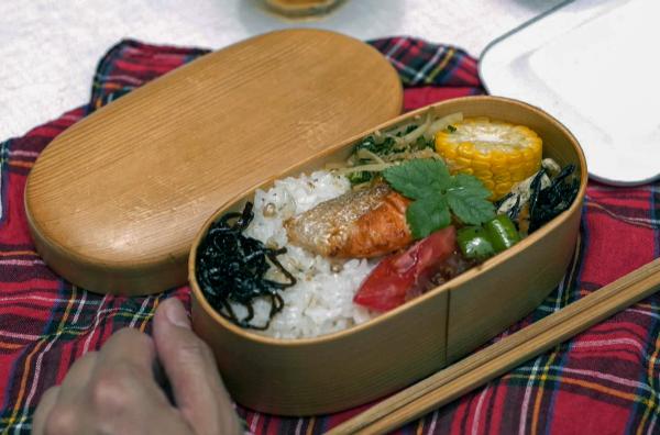 Bild 1 von 3: Eingelegtes Gemüse, gekochter Mais, Rettichstängel mit Sesam und Lachsscheiben werden für das Bento auf dem gekochten Reis verteilt.