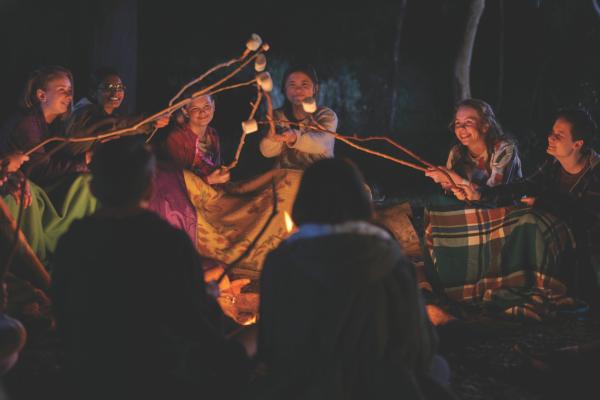 Bild 1 von 8: Natalie (Nandini Rajagopal, h.2.v.l.), Hannah (Evie MacDonald, h.3.v.l.), Olivia (Elena Liu, h.M.), Sarah (Jake Childs, h.r.) und andere Mitschülerinnen sitzen gemeinsam beim Lagerfeuer. Noch ahnt Hannah nicht, welche intimen Fragen einige von ihnen ihr stellen werden.