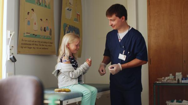 Bild 1 von 3: Der Arzt erklärt Vilda (Sofia Sittnikow), was beim Impfen passiert und lässt Vilda die Banane impfen. Danach darf er Vilda impfen.