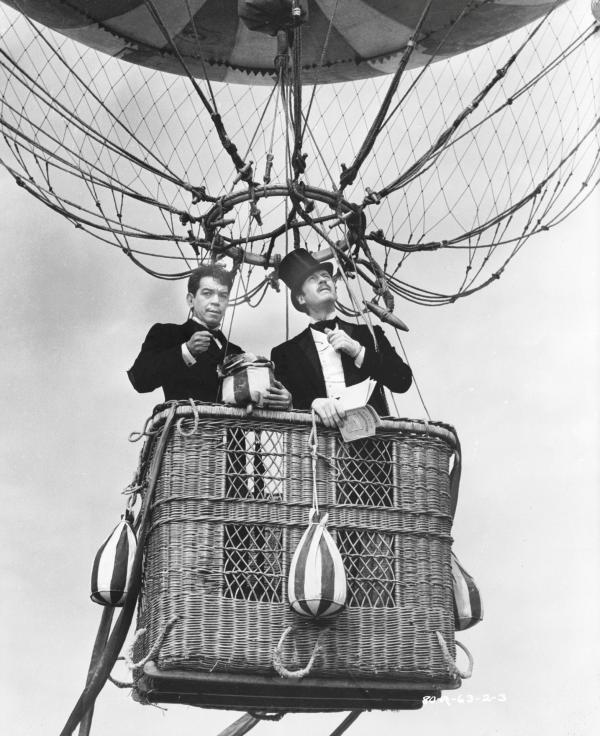 Bild 1 von 11: Full shot of Cantinflas as Passepartout and David Niven as Phileas Fogg, wearing top hat; both standing in basket of hot air balloon.