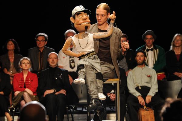 Bild 1 von 6: Mit seiner Puppe Herbert trat Simon Janetski (Paul Boche, M.) bei einem Wettbewerb gegen das Opfer an - genauso wie auch andere Kleinkünstler (Komparsen, Hintergrund).